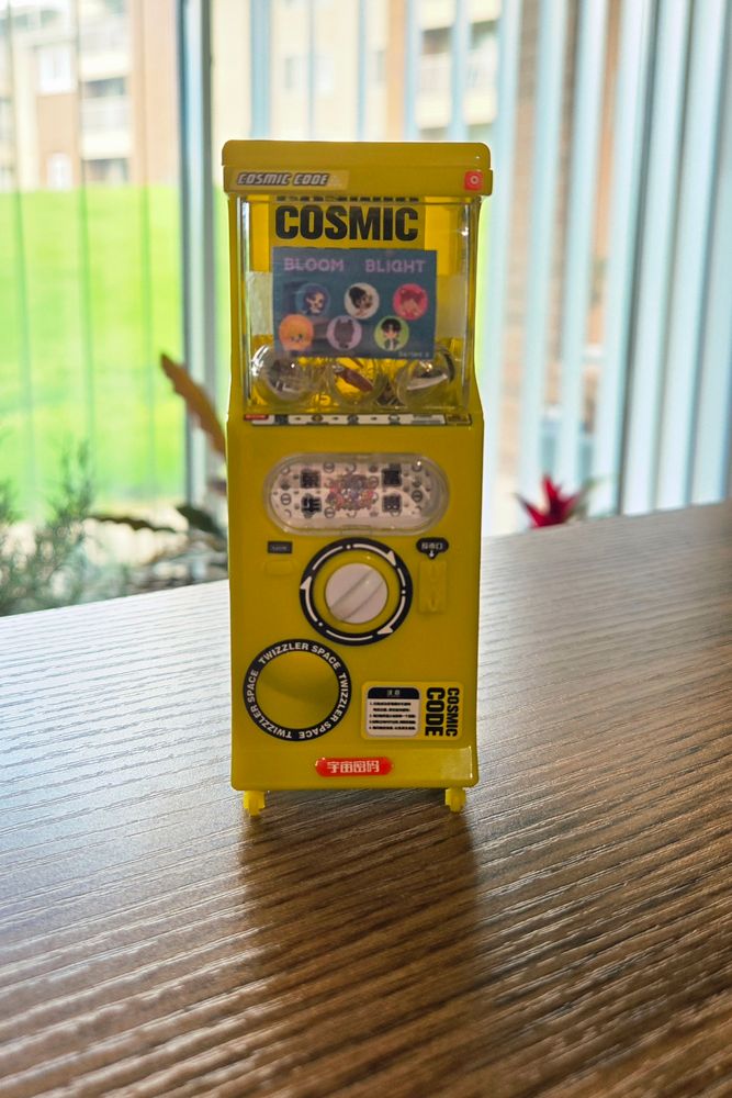 Photo of an itty bitty little yellow gachapon machine model sotting on a wood table in fro t of a window. Factory stickers all over read "Cosmic Code" or "Twizzler Space", but a handmade sign has been stuck inside the window showing Bloom&Blight themed prizes- each of the characters chibi art. The inside is full of clear capsules, each with one character's art in.