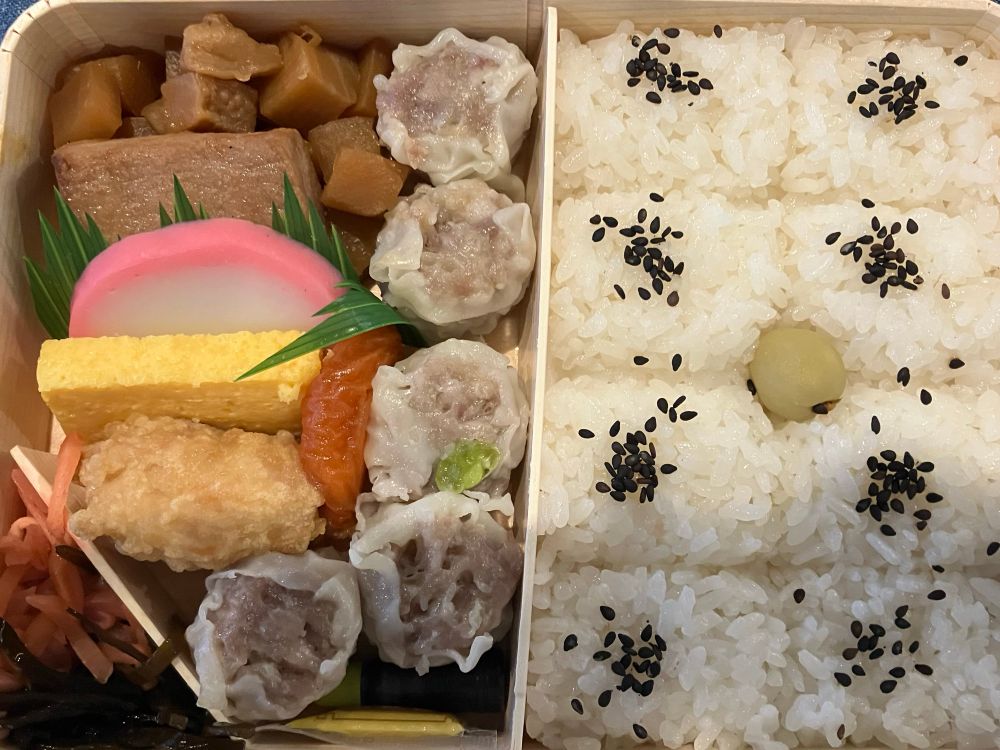 崎陽軒のシウマイ弁当の写真。左側がおかず、右側がご飯スペース。メインのシウマイ5個にたけのこ炒め煮と煮魚、唐揚げ、卵焼き、かまぼこ、ほしあんず、お漬物と昆布佃煮。ご飯は8等分に一口サイズに分けられていてそれぞれに胡麻のトッピング。真ん中に青ウメ。