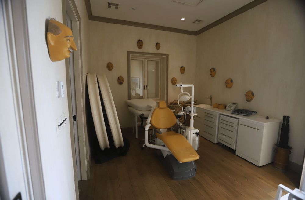 A room with a series of masks of men's faces on the walls and on a shelf with a yellow medical examining chair in the middle of the room.