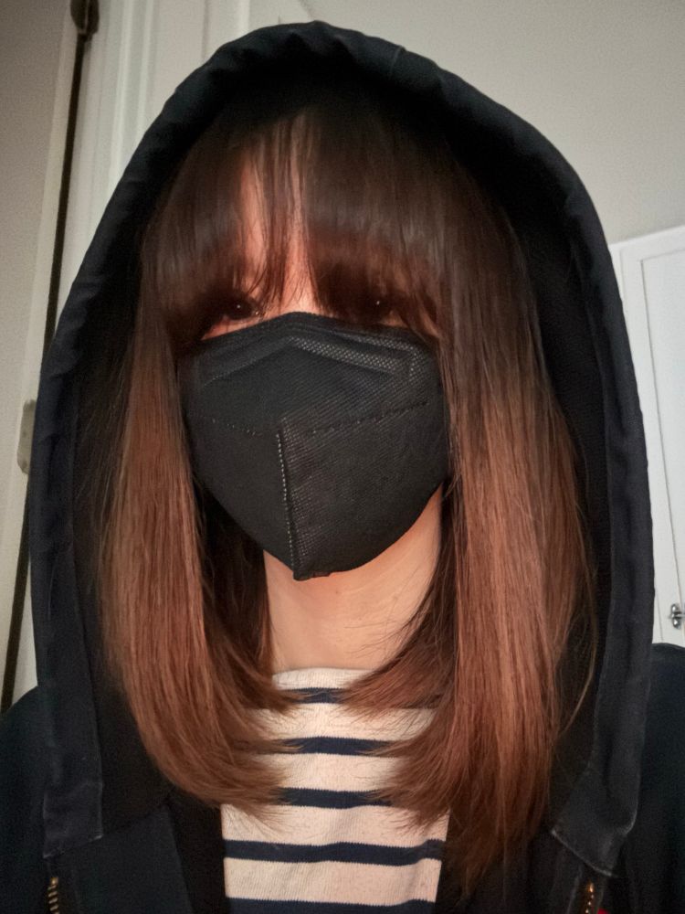 Photo of a girl with short brown hair and long bangs covering her eyes wearing a hood and face mask