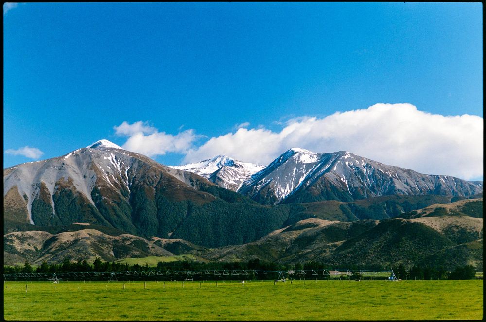 Photo of a mountain range with snow on their peaks. Blue sky with some white clouds behind the distant mountains. Green grass in the foreground - Fujifilm Superia Premium 400 - Pentax Super Program
