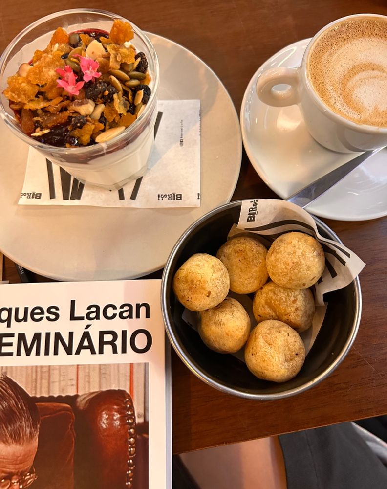 Vistos de cima sobre uma mesa: um parfait, um capuccino e pães de queijo e um livro