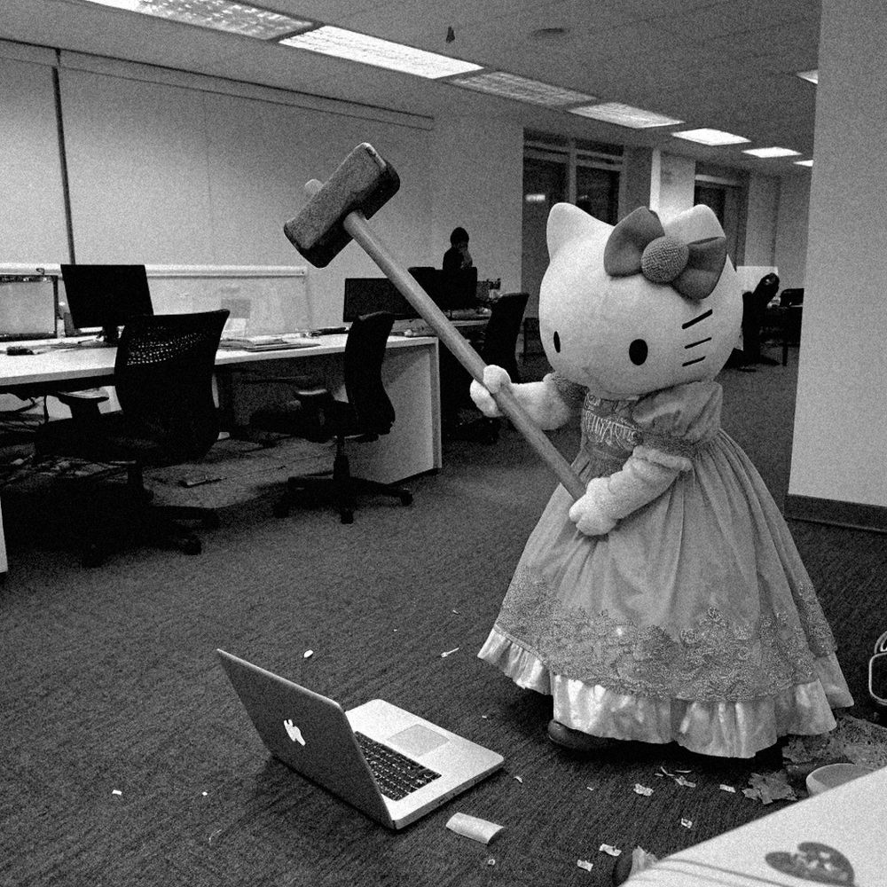 Uma hello kitty gigante erguendo uma marreta a ponto de marretar um MacBook que está no chão de carpete de um escritório.
