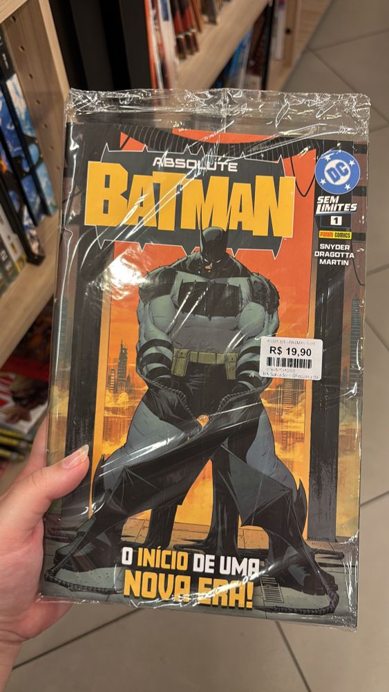 Foto de uma mão segurando o primeiro volume da HQ Absolute Batman. O Batman tem um porte físico mais cheio, no estilo “armário”, está sério, com a cabeça levemente abaixada, e segurando dois escudos, que também funcionam como ganchos, com forma de asas de morcego. 