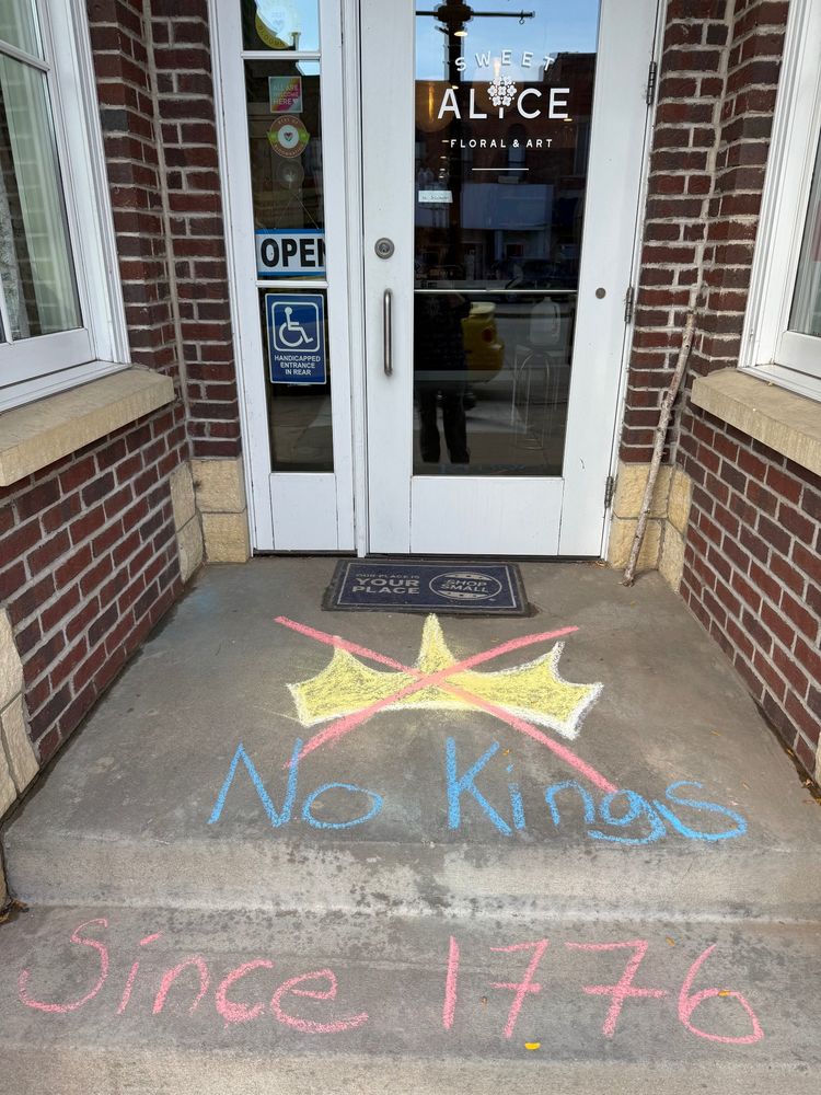 The front entrance to Sweet Alice Floral & Art is pictured, with chalk art reading “No Kings since 1776” below a drawing of a crown crossed out.
