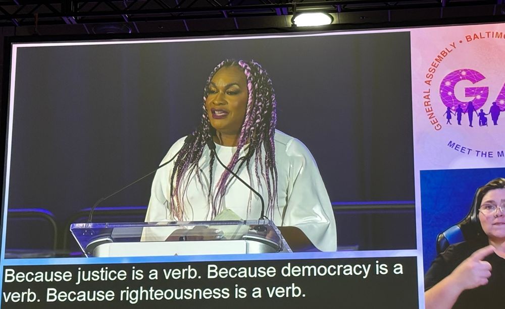 A tall, imposing, elegant black woman with black-and-pink extensions speaks at a podium.