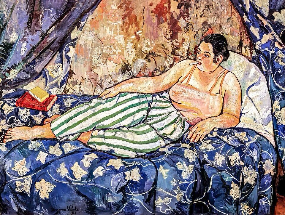 A painting of a heavyset woman reclining on a bed. She’s wearing what seem to be pajama pants and a camisole, and smoking a cigarette. There are some books by her side. She’s surrounded by lush, patterned fabrics. She’s not looking at the viewer. She’s thinking her own thoughts.