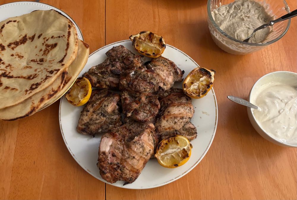 White serving dishes on a wooden table hold spice-rubbed grilled chicken things, grilled lemon halves, naan with char marks on it, and two bowls with puréed sauces - one off-white, one tan.