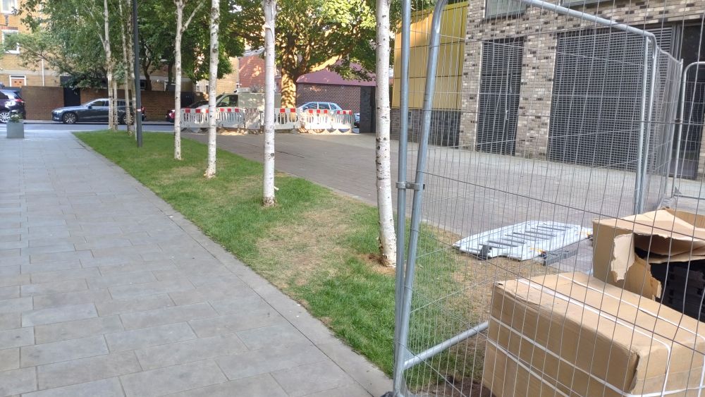 Two paved paths leading to a road in the distance. In between them is a patch of grass with trees in a line. Between the paved paths, over the grass, is a path where the grass has been worn out. In the foreground is a temporary fence where attempts are being made at encouraging people to walk a particular line. 