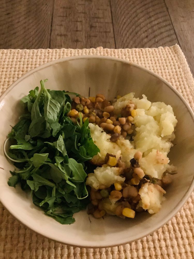 A wide bowl half full of torn spinach and arugula and half full of a mashed potatoes, beans, and mixed vegetables 