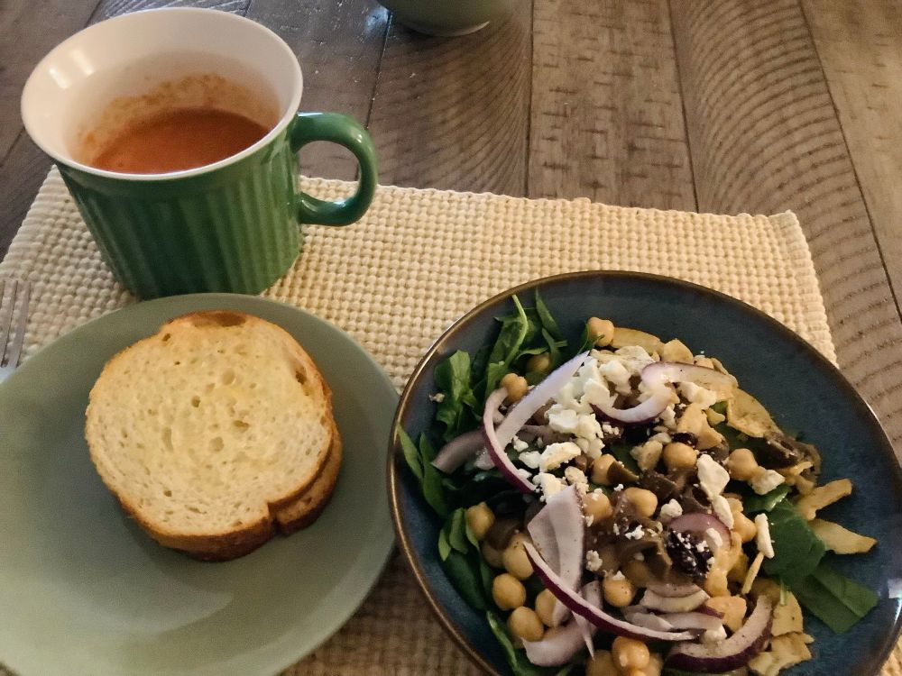 A grilled cheese sandwich on round bread on a plate next to a salad with spinach, garbanzo beans, red onions, feta, and olives. And there's a big mug of tomato soup.