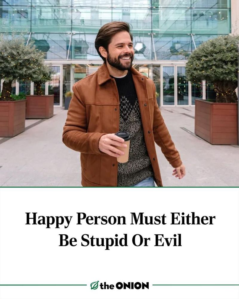 A meme from the ONION that says, "Happy Person Must Be Stupid Or Evil". The image shows a younger, bearded man smiling and appearing what is conventionally thought of as happy. He's wearing a light brown suede coat and a over sized sweater. He's holding a coffee to-go cup in his right hand and he appears to be standing in the well lit atrium or lobby of a large office building.