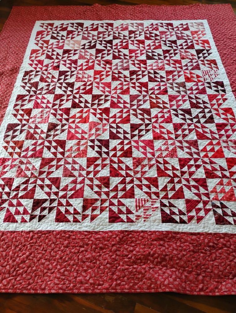 Huge (80" x90" approx) red and white quilt, using North Wind blocks and a wide variety of red prints. Blocks are made of a large red triangle in one corner, large white triangle in opposite corner, and red and 5 small triangles in each of red and white in between the large triangles. When set solid, but alternating which corner is red, they form interesting secondary patterns. Quilt also has a wide red paisley border.