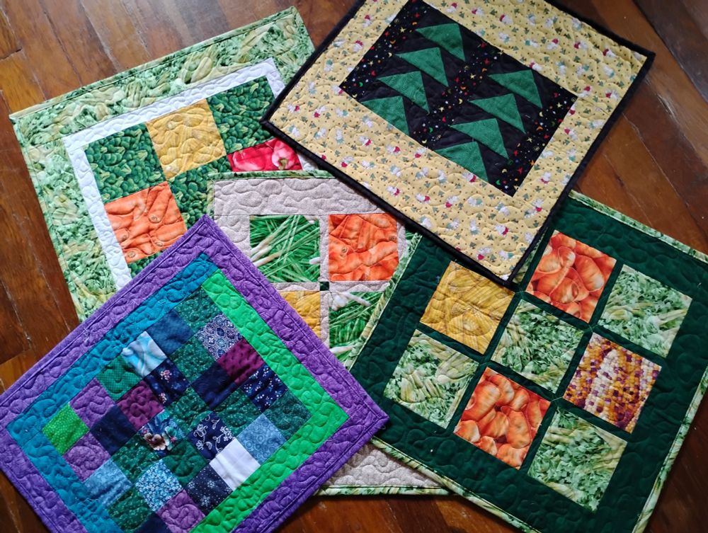 Five little quilts, about 18" square, three made with photo-realistic prints of vegetables (carrots, corn, broccoli, peppers, leeks, greens) in single, large nine-patch layouts, one made of random prints (purple, neon green, blue) cut in squares, and one made of green flying gees on a black background with an odd yellow print border that has teddy bears in red or green shorts and little Christmas trees!
