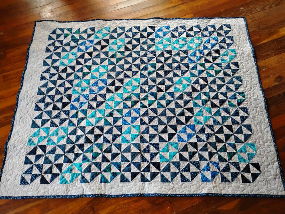 Broken Dishes blocks (alternating dark and light triangles) in dark blue and turquoise/teal on a mottled light blue background, set solid, so they have the same basic overall effect as pinwheel blocks, with a bit of mottled blue border and dark blue binding. The lighter colored turquoise blocks create secondary patterns when three or four are touching, either horizontally or diagonally.