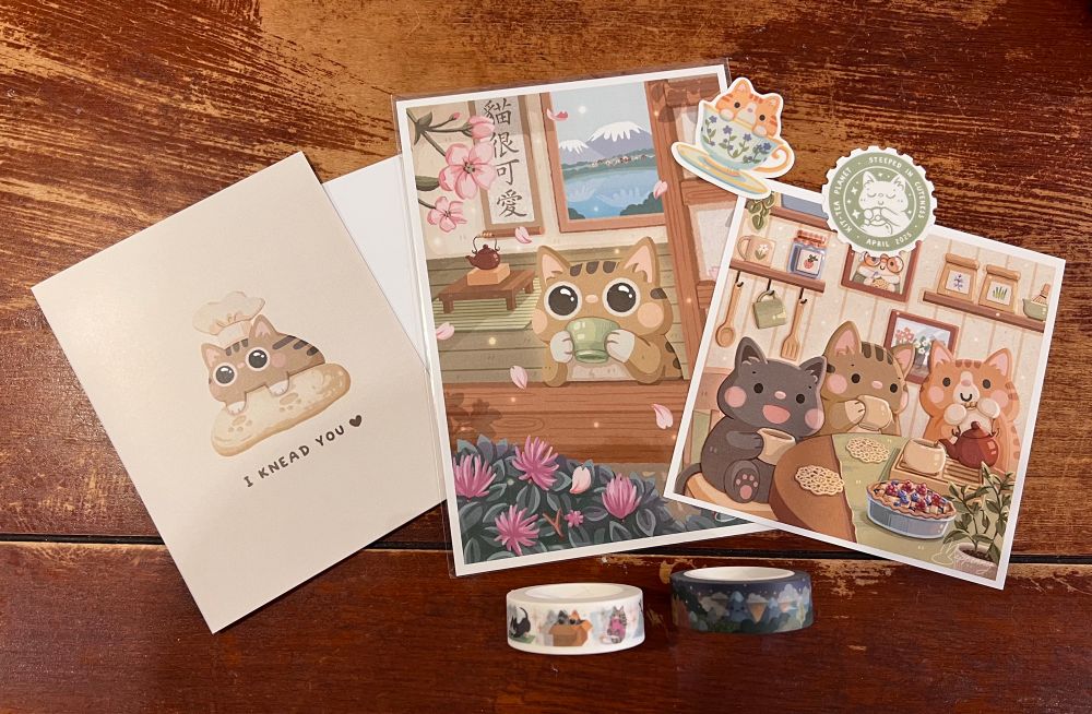 A spread of stationary items and prints. A cat  kneading bread on a card. A print of a cat at a window sipping tea. Three cats enjoying a tea party. Two washi tapes- one with cats and the other with a landscape. A sticker of a cat in a teacup and another sticker of a faux stamp of a cat.