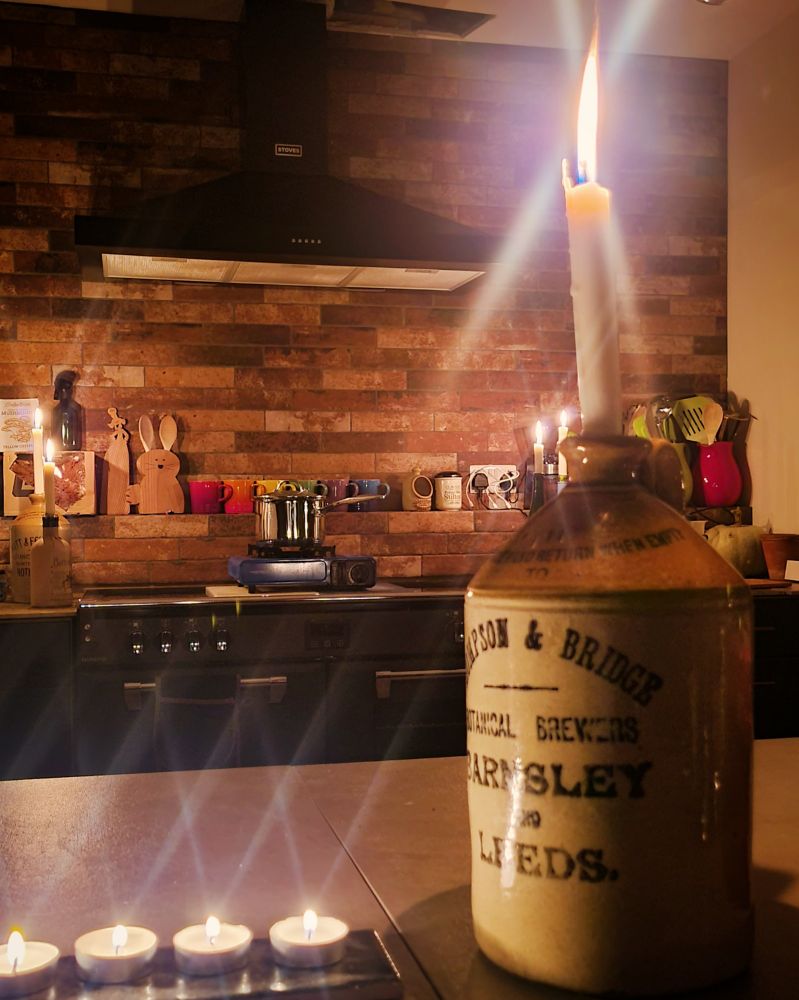 An old brewery keg with a candle in it in the foreground, a kitchen in candlelight behind.