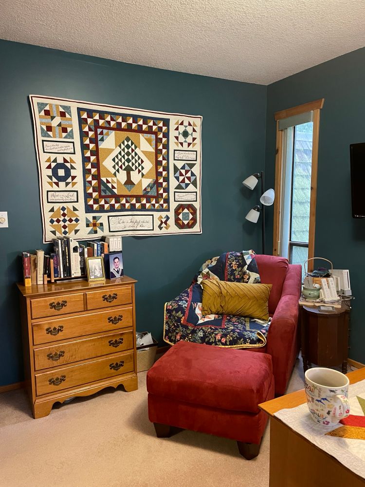 ‘Sophisticated Teal’ paint colour behind Sampler quilt I. Red, gold, teal on cream; red chair and ottoman with navy/red/gold/teal quilt draped over.