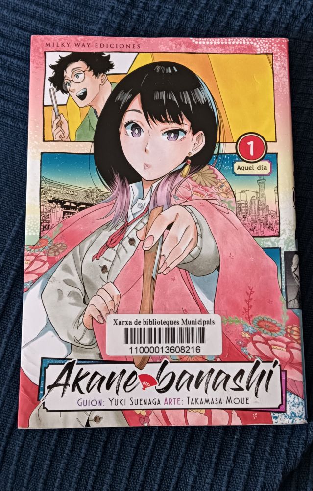 Sobre una tela azul oscuro, un tomo de manga:

Akane Banashi, tomo 1.

En la cubierta destaca la figura de una chica vestida de color rosa intenso, que sostiene delante de sí un abanico cerrado. Tiene el pelo largo y oscuro hasta los hombros y lleva las puntas teñidas de rosa. Tiene un gesto interrogativo.