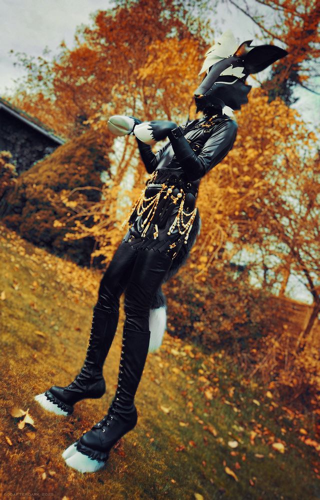 image is a photograph of a person dressed in pony play attire, themed with a mask resembling a monochromatic kirin. their outfit is accented with gold chains with white pearls and black tassels. behind them is a wall of golden autumn trees. #fursuit #petsuit