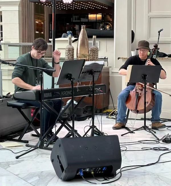 Sudowoodo Duo playing a set at Belvedere lobby bar at Super MAGFest. Alex carefully reads sheet music with his cello in hand while Alex engages the crowd with his keys, captured mid medley