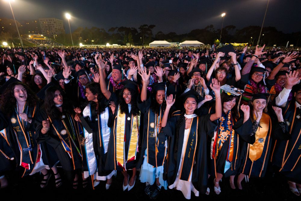 A group of joyful graduates in caps and gowns, celebrating at an outdoor UC San Diego commencement at night. Some are waving their hands and smiling broadly.