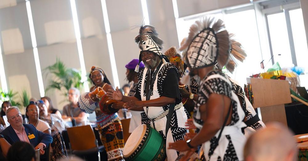 Performers in cultural attire playing drums and dancing at an indoor event