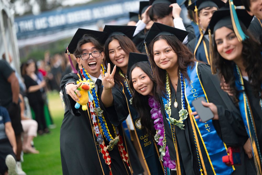 Group of joyful graduates in caps and gowns celebrating, with some wearing leis and holding diplomas, at an outdoor graduation ceremony.
