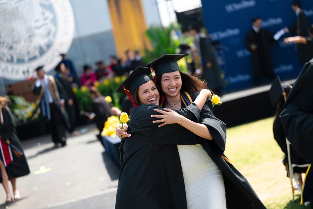 Two graduates hug joyfully at their commencement ceremony, holding yellow roses and wearing black caps and gowns.