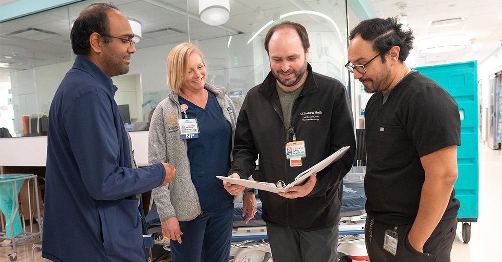 Four medical professionals are engaged in a discussion around a clipboard in a hospital corridor.