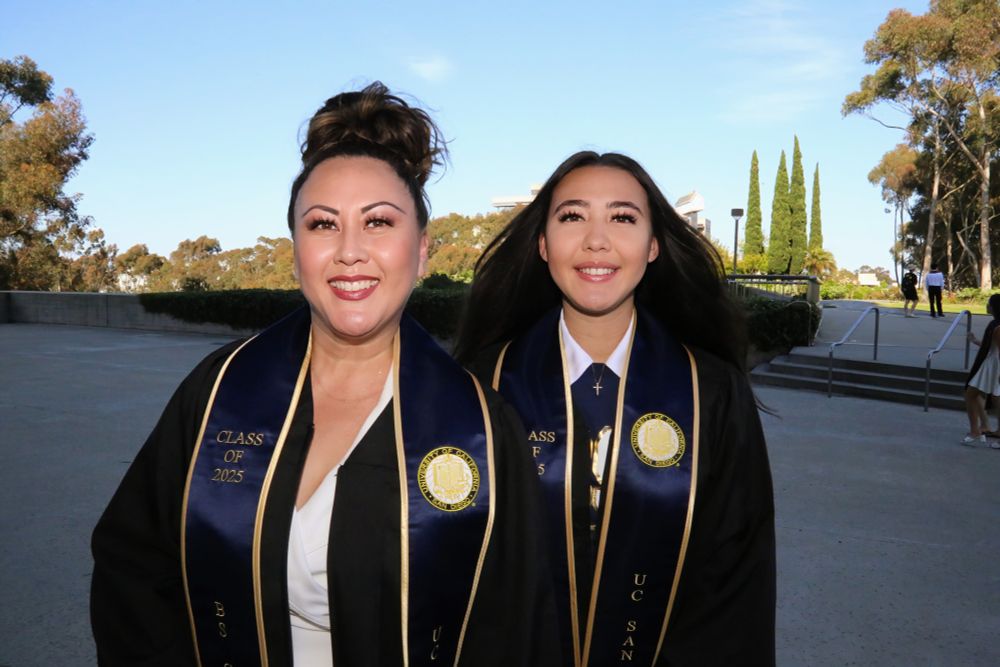 Two graduate smiling, wearing sashes and UC San Diego regalia, standing outdoors on a sunny day.