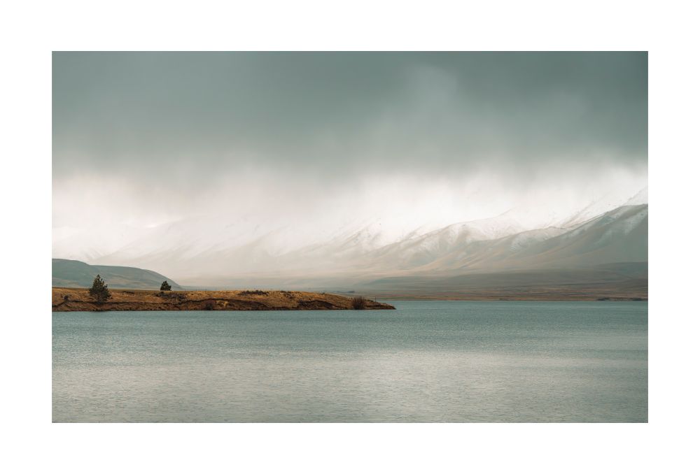 A man made lake from the 1930s with a headland & 2 trees , a mountain range in the distance with snow on it & heavy clouds dropping light snow