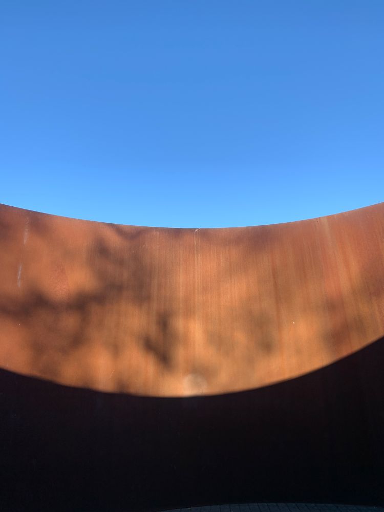 A large curved, rusted steel wall with mottled sunlight in front of a bright blue sky. It is part of a large Richard Serra sculpture
