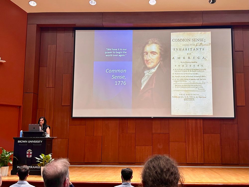 A projector screen in front of a wooden wall on a state with a woman with dark hair in a black shirt standing behind a podium 