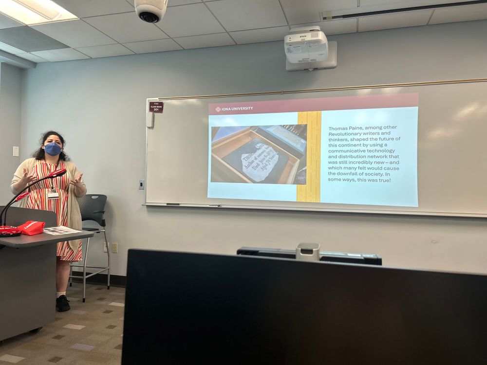 A woman in a striped desk and face mask presents in front of a screen with an image of a grave stone and text