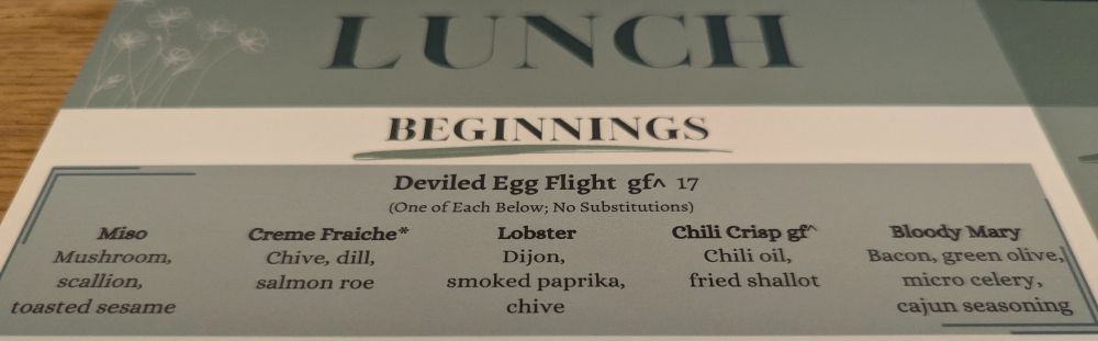 Olive green and creme colored menu, with white outlines of poppies in the upper left. The menu reads: 
LUNCH

BEGINNINGS

Deviled Egg Flight gf^ 17

(One of Each Below; No Substitutions)

Miso
Mushroom, scallion,
toasted sesame

Creme Fraiche*
Chive, dill,
salmon rое

Lobster 
Dijon, smoked paprika, chive

Chili Crisp gf^
Chili oil,
fried shallot

Bloody Mary
Bacon, green olive, micro celery,
cajun seasoning