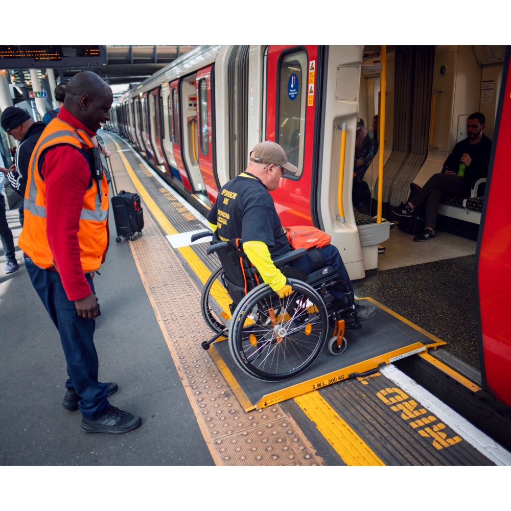 Me in my wheelchair being helped by TfL staff ramp assistance to board a tube train. A member of staff is standing behind me watching out to make sure I board safely while im moving across the ramp and into the tube car between the open doors.