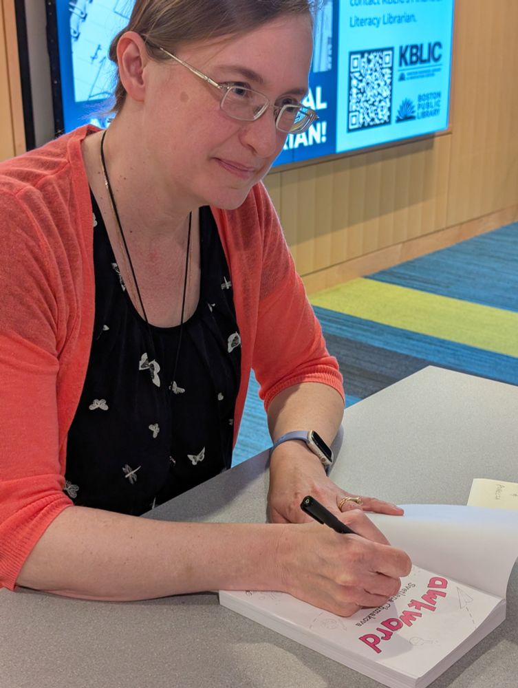 An image of Svetlana Chmakova, a woman wearing glasses, signing a copy of Awkward and looking at an attendee. 