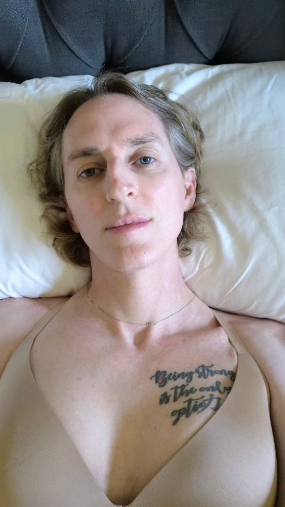 Image of a nonbinary person laying on a bed with white sheets and a dark gray headboard. They're in a beige bra and their appearance is soft and sleepy.