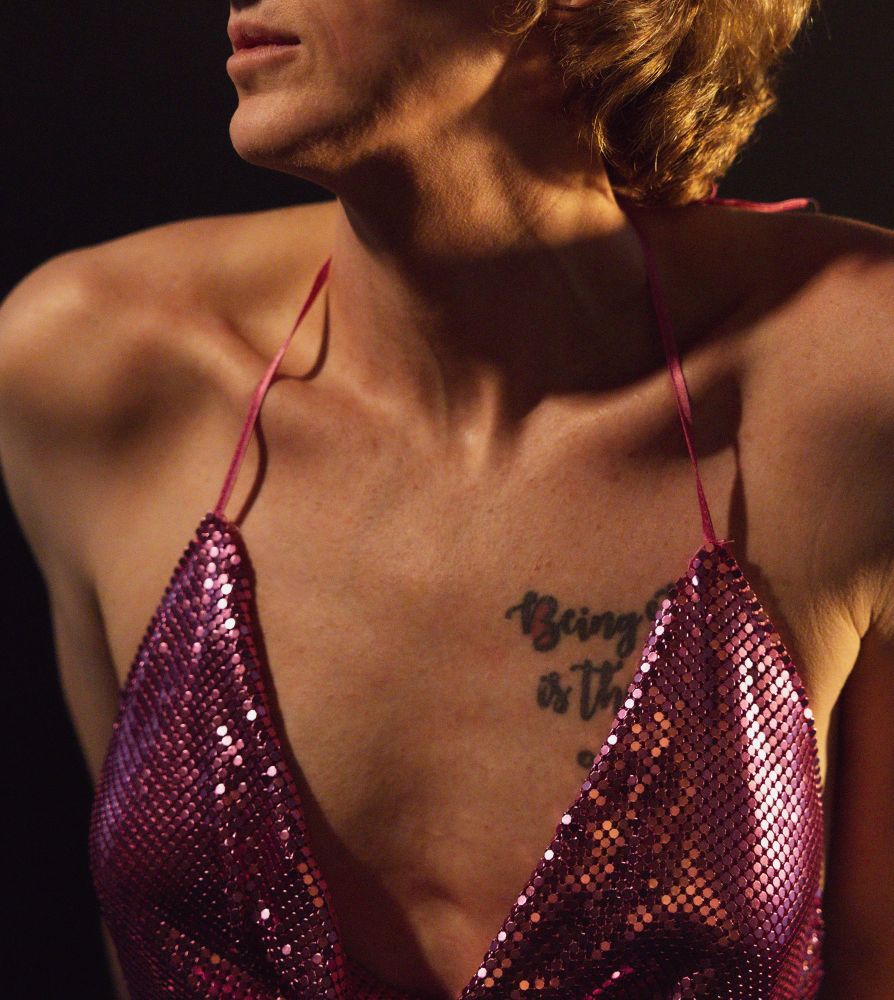 A close-up of a person wearing a sparkling pink outfit with thin straps. Part of a tattoo that reads "Being" is visible on the chest. The background is dark, creating a dramatic effect.