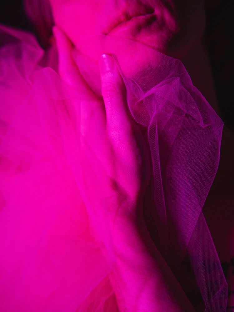 A close-up image of a person's hand gently touching a translucent, pink fabric, with dramatic pink lighting creating soft shadows on the skin and fabric. The overall mood is intimate and artistic.