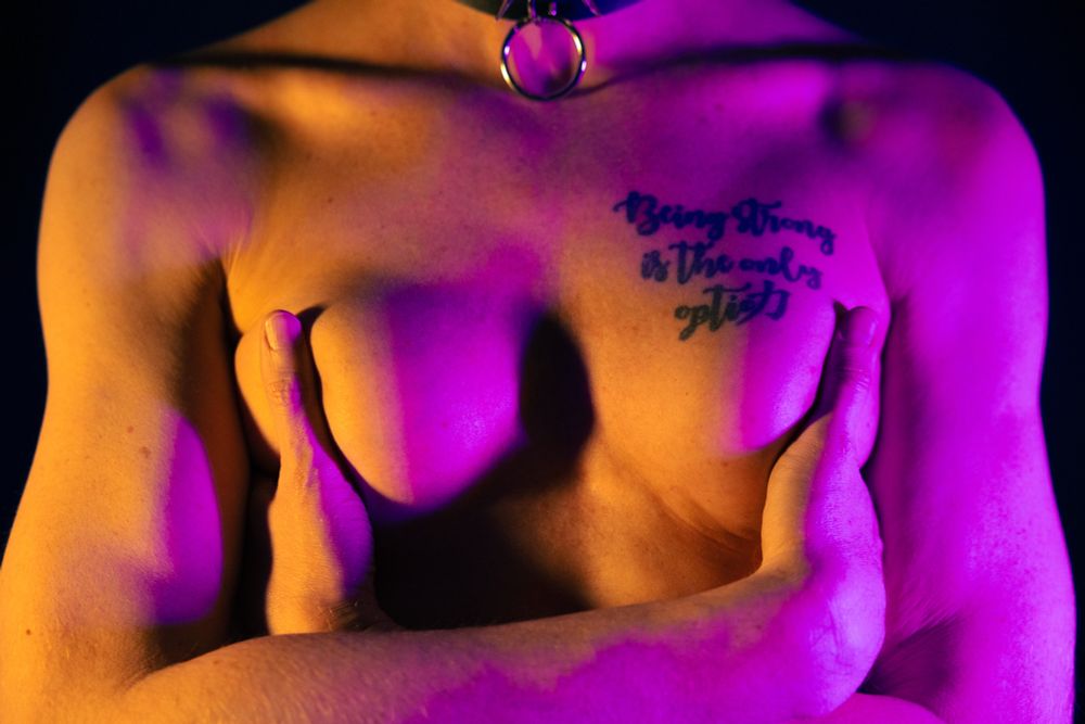 The image features a close-up of a person's upper body, showcasing bare skin with a visible tattoo that reads "Being strong is the only option." The individual is adorned with a choker, and the scene is illuminated with purple and blue lighting, creating a dramatic effect.