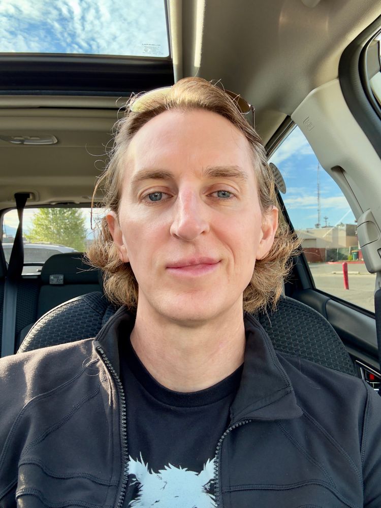 Image of a nonbinary person in a rental car, their hair is held back by a pair of cute aviator sunglasses, the sun shining through the car windows and sunroof. They’re in all black and sporting a half smile that says “I will top you”.