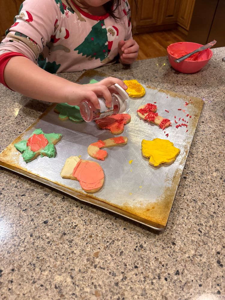 Person in pink Christmas pajamas decorating cookies with red, green, and/or yellow frosting. 