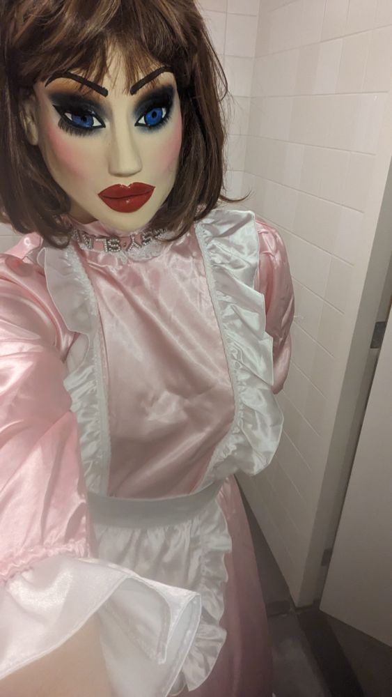 Sissy Holly wearing her long pink maids dress with white apron. She's also wearing her female mask and a dark brown beehive style wig
