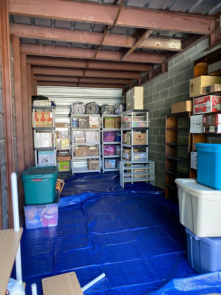 Photo of a storage unit, lined with blue tarps on the bottom and multiple storage shelves filled with boxes and bins. A broken shelf is in the foreground. 