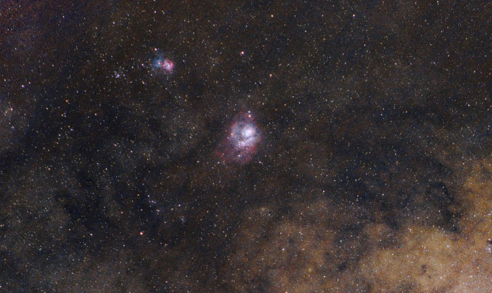 Picture of a region of sky around the Lagoon Nebula (near centre) and the Trifid Nebula (above and to the left of the Lagoon), both in Sagittarius. The Lagoon Nebula is seen in mixing shades of pink, cyan, and white, while the Trifid has two distinct sections, one in magenta and one in blue/cyan. The Trifid's magenta section, an emission nebula, has dust filaments that appear to cut it into thirds, giving it its name. Its blue/cyan portion is a reflection nebula.
The field covers an area approximately 6.5 degrees wide by 3.7 degrees tall. A few open and globular clusters can also be seen in the image. A bright area containing dust, gas, and stars from the central region of the Milky Way is visible at the bottom right.