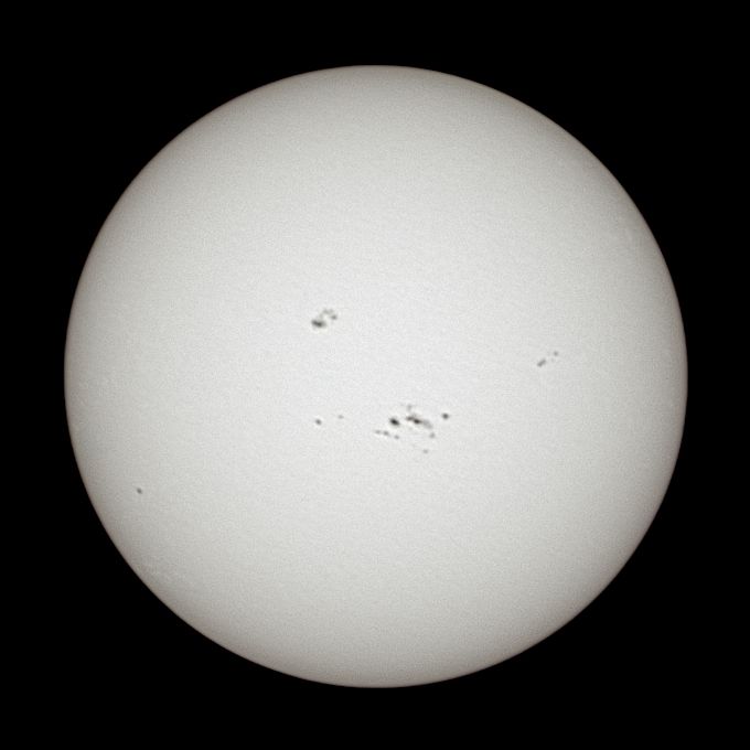 Photo of the Sun, seen as a white orb with a gentle texture to it and various spots. Active Region (AR) 4100 is visible just below the centre of the disk with 26 spots, and AR 4099 is just above and to the left of centre with 13 spots.
ARs 4096 (bottom left), 4101 (left of 4100), and 4104 (above+right of 4100) are also visible, with 1-3 spots each.
North is approximately down.