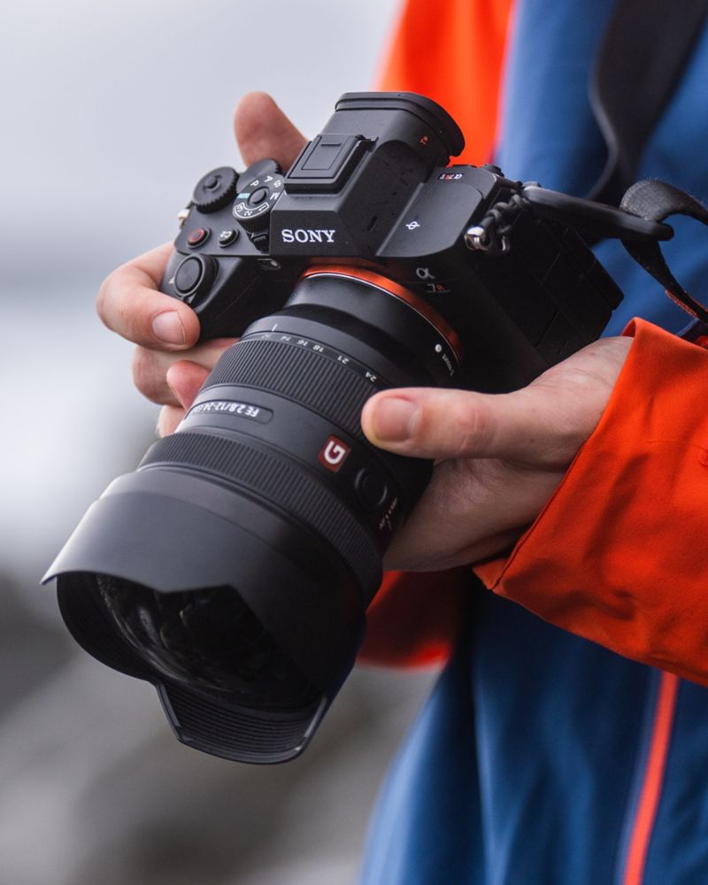 Photo of someone wearing a blue and orange jacket holding a professional Sony camera with both hands. The background of the photo is blurred, and the camera focuses on the person's hands and the Sony camera. 