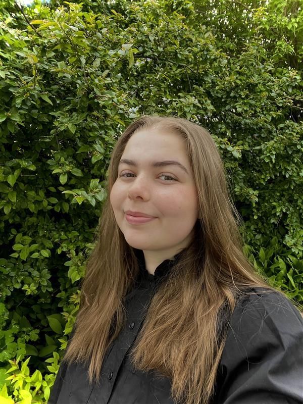 Portrait photo of a woman smiling with long, light brown hair and wearing a black shirt, standing in front of green foliage.